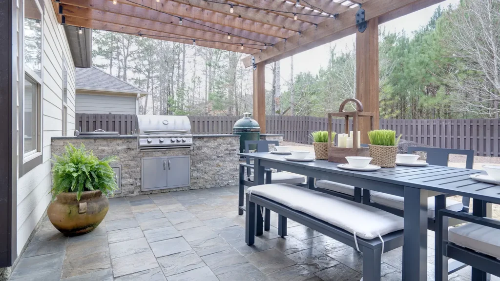 Pretty summer outdoor kitchen with table set and grilling station underneath wooden arbor on stone patio.